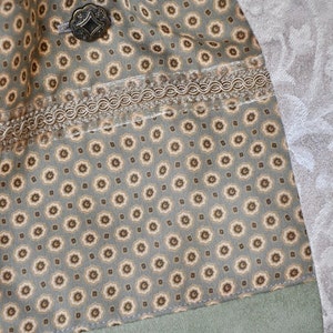 Puede incluir: Primer plano de una tela verde con un patr&oacute;n floral marr&oacute;n y blanco. La tela tiene un ribete marr&oacute;n y una trenza decorativa. Un bot&oacute;n de metal es visible en la esquina superior izquierda.