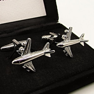 以下が含まれることがあります： 黒いベルベットの箱に入った銀色の飛行機のカフスボタンのペア。箱の蓋には「Style Haus」の文字が書かれています。