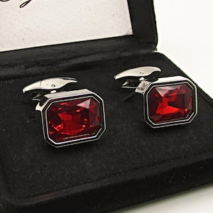 Puede incluir: Un par de gemelos con grandes piedras preciosas rojas rectangulares engastadas en metal plateado. Los gemelos se presentan en una caja forrada de terciopelo negro. La caja tiene el texto "Style Haus" en el interior.