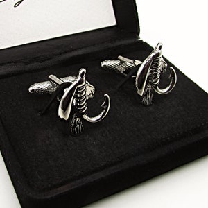 Puede incluir: Gemelos plateados con diseño de mosca de pesca, presentados en una caja negra. La caja abierta revela un interior blanco con el texto "Style Haus" en escritura elegante. Un accesorio elegante.