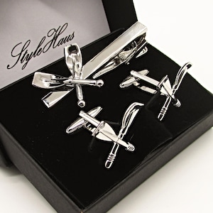 May include: A silver tie clip and cufflinks set in a black box. The tie clip and cufflinks feature a crossed oar design. The box has the words "Style Haus" printed on the inside lid.