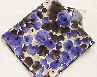 Può includere: Un fazzoletto da taschino a fantasia floreale, con fiori blu e marroni su sfondo crema. Il fazzoletto è piegato a forma di diamante, con un bordo cucito bianco. Il tessuto sembra essere cotone o simile.