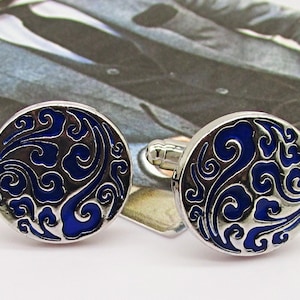 Puede incluir: Un par de gemelos plateados con un diseño circular. Cada gemelo presenta un patrón azul oscuro y arremolinado sobre un fondo plateado. Los gemelos se colocan sobre un fondo borroso de un traje.