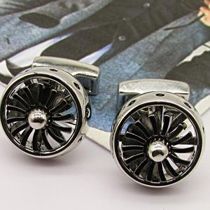 Puede incluir: Un par de gemelos plateados con un diseño de motor de turbina. Los gemelos presentan un patrón detallado de aspas de ventilador dentro de un marco circular, creando una estética mecánica. El fondo es una imagen borrosa de una persona con traje.