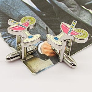 May include: A pair of silver cufflinks featuring a cocktail design. Each cufflink has a martini glass shape with pink, yellow, and blue enamel details. The cufflinks are presented on a patterned background.
