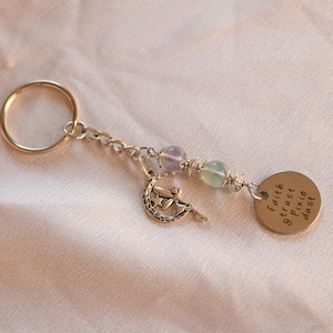 Puede incluir: Un llavero plateado con un dije de un hada en una luna creciente, cuentas verdes y moradas, y una etiqueta redonda plateada con las palabras "faith trust pixie dust" grabadas.