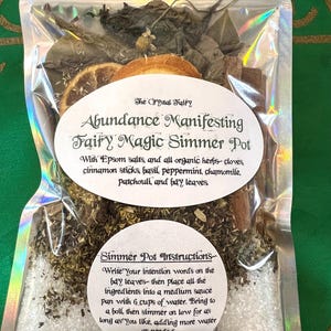 Puede incluir: Una bolsa de plástico transparente llena de hierbas secas, especias y sales de Epsom. La bolsa está etiquetada como "Abundance Manifesting Fairy Magic Simmer Pot" e incluye instrucciones de uso. Los ingredientes incluyen hojas de laurel, palitos de canela, albahaca, menta, manzanilla, pachulí y sales de Epsom.