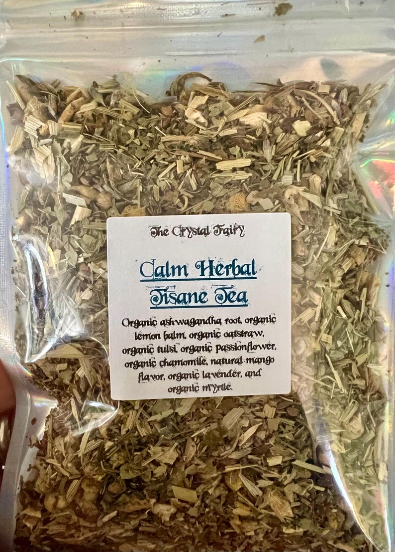 May include: A clear, iridescent bag filled with dried herbs and tea leaves. A white label reads "The Crystal Fairy" and "Calm Herbal Tisane Tea." Ingredients include ashwagandha root, lemon balm, oatstraw, tulsi, passionflower, chamomile, mango flavor, lavender, and myrtle.
