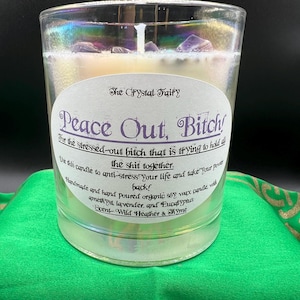 May include: A clear glass candle with a white label that reads "Peace Out, Bitch!" and "The Crystal Fairy." The candle contains purple crystals and is on a green surface. The candle is handmade with organic soy wax.