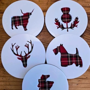 Könnte beinhalten: Set aus fünf weißen Untersetzern mit schottischen Motiven. Jeder Untersetzer hat einen anderen Ausschnitt, der mit rot-schwarz kariertem Stoff gefüllt ist. Zu den Designs gehören ein Highland-Rind, eine Distel, ein Hirsch, ein Scottish Terrier und eine Karte von Schottland.