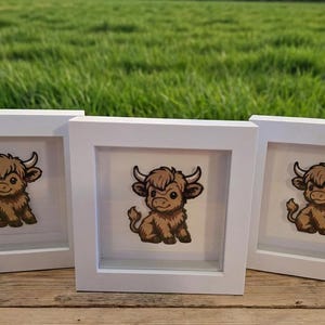 Op de afbeelding: Drie ingelijste prints met een cartoon Highland-koe. Elke print toont een bruine en bruine koe met grote hoorns, tegen een witte achtergrond in een witte vierkante lijst. De lijsten staan op een houten oppervlak.