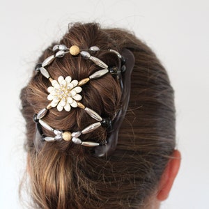 Puede incluir: Una pinza para el pelo marrón con cuentas plateadas y negras y una flor blanca con pedrería. La pinza sujeta el pelo marrón de una mujer en un moño.