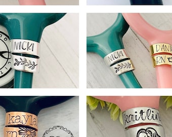 Könnte beinhalten: Sechs personalisierte Stethoskop-Anhänger mit verschiedenen Namen und Designs. Die Anhänger sind aus Metall und haben eine silberne, goldene oder kupferne Oberfläche. Die Designs beinhalten einen Herzschlag, eine Krone, eine Blume und einen Zweig.