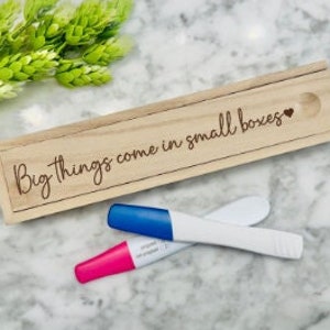 Puede incluir: Una caja de madera con el texto "Big things come in small boxes" grabado en ella. Dos pruebas de embarazo están sobre una superficie blanca junto a la caja.
