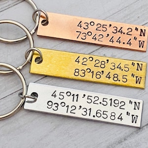 May include: Three silver keychains with engraved coordinates. The top keychain is copper, the middle keychain is gold, and the bottom keychain is silver. The coordinates are 43°25′34.2"N 73°42′44.4"W, 42°28′34.5"N 83°16'48.5 "W, and 45°11'52.5192 "N 93°12′31.6584"W.