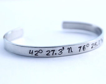 May include: A silver-colored cuff bracelet with engraved coordinates: 42° 27.3' n 76° 25.1' w. The open-ended bracelet is made of a shiny, metallic material and is set against a white background. The engraved text is in black.