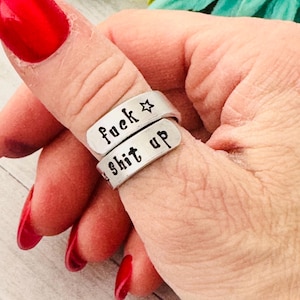 May include: Silver-toned wrap ring with the words "fuck" and "shit up" stamped on it. The word "fuck" has a star symbol next to it. The ring is worn on a finger with red nail polish.