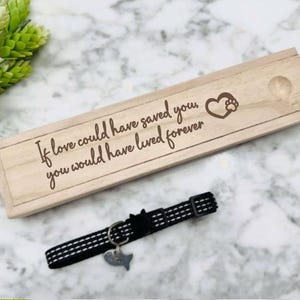 May include: A wooden box with the inscription "If love could have saved you, you would have lived forever."  A black and white dog collar with a paw print charm is in front of the box.
