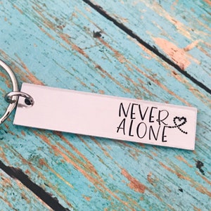 May include: Silver keychain with a rectangular tag that reads "NEVER ALONE" with a heart and chain design.