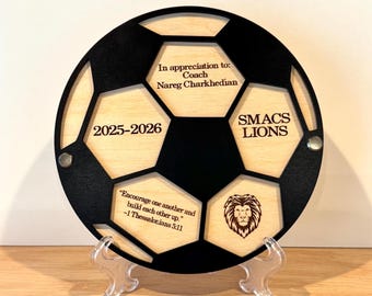 May include: A wooden soccer ball-shaped plaque with black and natural wood tones. The plaque includes text that reads "In appreciation to: Coach Nareg Charkhedian" and "SMACS LIONS". It also features the years "2025-2026" and a quote.