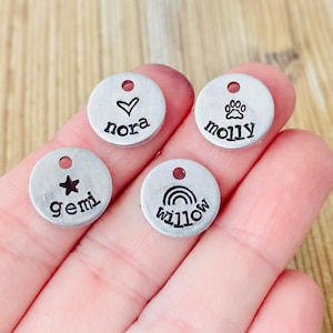May include: Four silver metal tags with different designs and names. The tags have a hole at the top for attaching to a keychain or necklace. The tags feature a heart with the name "nora", a paw print with the name "molly", a star with the name "gemi", and a rainbow with the name "willow".
