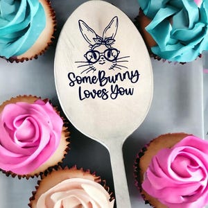 May include: A silver spoon with a bunny face and the text "Some Bunny Loves You" is surrounded by cupcakes with pink and blue frosting. The cupcakes have a light brown base and are arranged around the spoon.