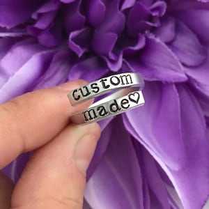 May include: Silver ring with the words "custom made" and a heart engraved on it. The ring is on a finger against a purple flower background.