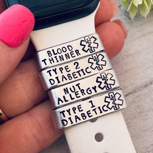 May include: Silver medical alert charms for a smartwatch band. The charms are stamped with the following conditions: Blood Thinner, Type 2 Diabetic, Nut Allergy, and Type 1 Diabetic. Each charm has a medical symbol.