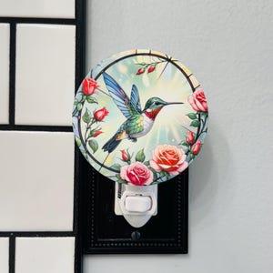 May include: A round night light featuring a colorful hummingbird with outstretched wings, surrounded by pink and red roses. The night light is set against a backdrop of a soft, glowing light and is framed by a black border.