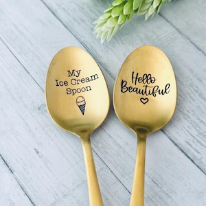 May include: Two gold-colored ice cream spoons. One spoon has the text "My Ice Cream Spoon" with an ice cream cone graphic. The other spoon has the text "Hello Beautiful" with a heart graphic. The spoons are on a light wood surface.