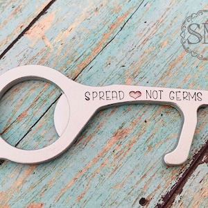 May include: Silver-toned key-shaped tool with a loop at the top and a small heart engraved on the handle. The tool is engraved with the text "Spread ♥ Not Germs".