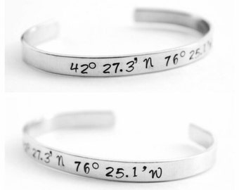 May include: Two silver bracelets with engraved coordinates. The top bracelet reads "42° 27.3' N 76° 25.1'" and the bottom bracelet reads "27.3' N 76° 25.1' W".