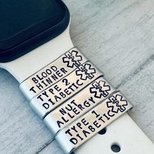 以下が含まれることがあります： 「BLOOD THINNER」「TYPE 2 DIABETIC」「NUT ALLERGY」「TYPE 1 DIABETIC」の黒い文字が入ったシルバーの医療アラートチャーム。各チャームには医療シンボルがあり、白い時計バンドに取り付けられています。