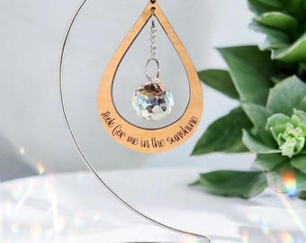 May include: A wooden teardrop-shaped ornament with the text "look for me in the sunshine" engraved on it. A clear crystal ball hangs inside the wooden frame. The ornament is suspended from a silver-colored metal stand.