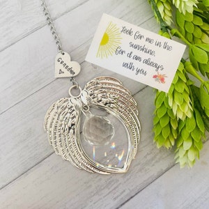 May include: A silver heart-shaped wind chime with angel wings and a crystal prism. The heart has the word "Grandma" engraved on it. A small card with the text "Look for me in the sunshine. For I am always with you" is in the background.