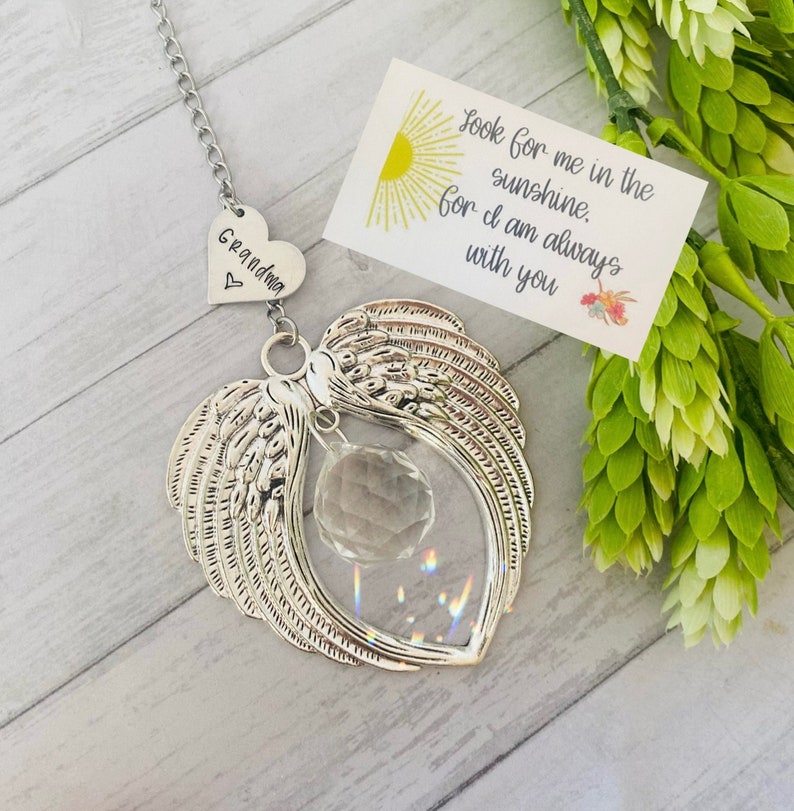 May include: A silver heart-shaped wind chime with angel wings and a crystal prism. The heart has the word "Grandma" engraved on it. A small card with the text "Look for me in the sunshine. For I am always with you" is in the background.