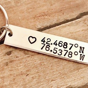 May include: Silver keychain with a heart and coordinates engraved on it. The coordinates are 42.4687° N 76.5378° W.