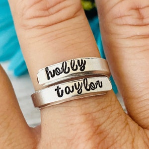 May include: A silver ring with the names "holly" and "taylor" engraved on it. The ring is worn on a finger.