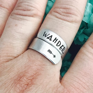 以下が含まれることがあります： 「WANDER」の文字が刻印されたシルバーカラーのラップリング。矢印が単語の下に刻印されています。指にはめられたリング。