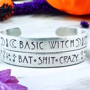 May include: Two silver metal cuff bracelets. The top bracelet reads "BASIC WITCH" with moon and exclamation point symbols. The bottom bracelet reads "BAT SHIT CRAZY" with star symbols. The bracelets are on a white surface.