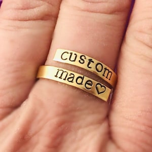 May include: A gold-colored wrap ring with the words "custom" and "made" stamped on it. The word "made" includes a heart symbol. The ring is worn on a finger.