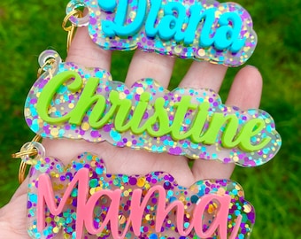 May include: Three personalized keychains with names "Diana", "Christine", and "Mama". Each keychain features a clear base filled with glitter, a colorful name, and a gold-colored keyring. The names are in blue, green, and pink.