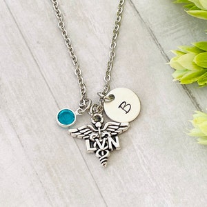 May include: Silver necklace with a medical caduceus charm, a blue crystal, and a silver charm with the letter "B".