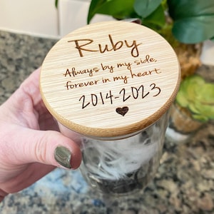May include: A clear glass jar with a bamboo lid, containing feathers. The lid is engraved with the name "Ruby", the years "2014-2023", and the phrase "Always by my side, forever in my heart" with a heart symbol.