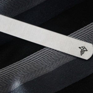 May include: Silver tie clip with a medical symbol engraved on it. The tie clip is on a black and white striped fabric.