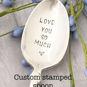 May include: A silver spoon with the words "LOVE YOU SO MUCH" stamped on the bowl, along with a small heart symbol. The text "Custom stamped spoon" is below the spoon. The spoon is surrounded by blue floral accents.