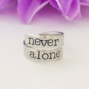 May include: A silver ring with the words "never alone" engraved on it. The ring is wrapped around a finger.