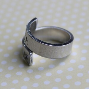May include: A silver ring with the word "Faith" engraved on the band. The ring is made of metal and has a simple design.