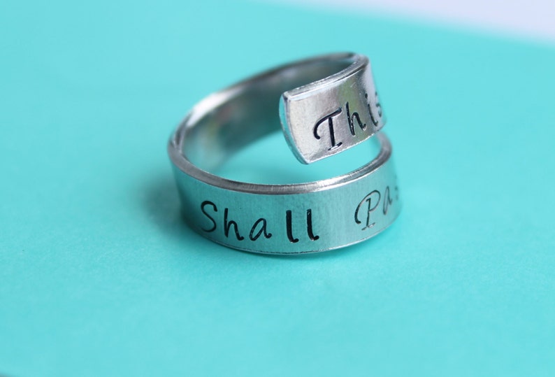 May include: A silver ring with the words "This Shall Pass" engraved on it. The ring is a twist design with the words wrapping around the band.