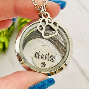 May include: Silver locket necklace with a paw print charm. The locket contains a small silver disc with the name "Charley" engraved on it and a paw print. The locket is filled with dark hair.